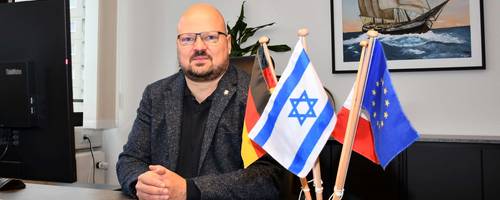 Bürgermeister Kai Eggert sitzt ernst an seinem Schreibtisch, auf dem ein Tischflaggenset mit der israelischen Flagge steht.