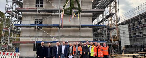 Eine größere Gruppe Menschen stehen vor einem mehrstöckigen Rohbau, über ihnen hängt der Richtkranz an einem Kran.