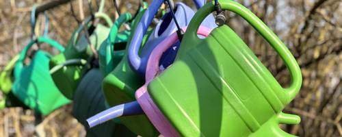 Eine Reihe bunter Gießkannen hängt nebeneinander. Alle Kannen sind mit Schössern angebunden.