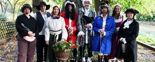 Eine Gruppe von Männern und Frauen stehen auf einem Platz vor einem großen Stein unter einem Blätterdach.