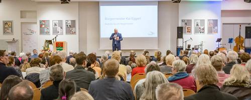 Blick über sitzende Menschen auf einen Redner an einem Rednerpult auf einer Bühne.