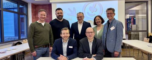 Zwei Männer sitzen an einem Tisch vor einer Projektion; hinter ihnen stehen fünf weitere Personen und schauen freundlich in die Kamera.