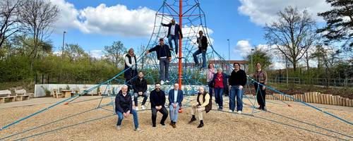 Eine Gruppe von 13 Personen hat eine Seilpyramide auf einem Spielplatz erklommen und sie schauen freundlich in die Kamera.