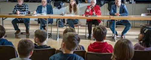 Ein Tisch, an dem mehrere Personen sitzen. Vor der Tischreihe befinden sich Kinder im Publikum. Von den Kindern sind nur die Rücken zu sehen.