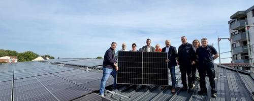 Eine Gruppe steht auf einer Dachfläche mit zahlreichen PV-Modulen und hält ein Solarpaneel in die Kamera.