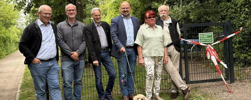 Eine Gruppe Männer und Frauen stehen mit einem Hund an der Leine vor einem eingezäunten Grundstück mit Wiese und Bäumen.