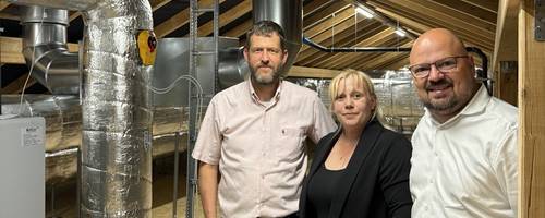 Drei Personen stehen in einem Dachgeschoss auf OSB-Platten und vor den Dachhölzern