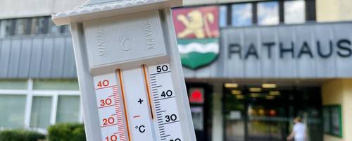 Eine Hand hält ein analogen Thermometer; im Hintergrund sieht man das Rathaus