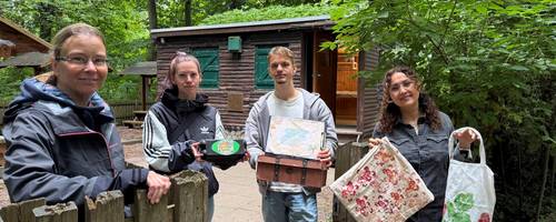 Drei Frauen und ein Mann stehen an einer Hütte im Wald und halten verschiedene Bastelwerke in die Kamera