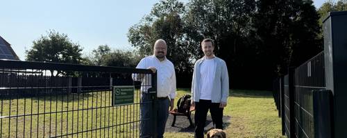 Bürgermeister Kai Eggert und Leiter des städtischen Teams Grünflächen stehen an einem geöffneten Tor vor einer Hundewiese. Davor sitzt ein mittelgroßer Hund mit braunem Fell..