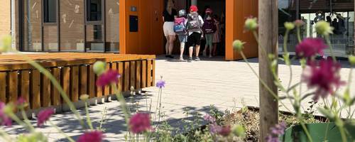 Grundschülerinnen und -schüler betreten mit Schulranzen auf dem Rücken einen orangenen Eingangsbereich einer Schule.