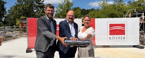 Zwei Männer und eine Frau halten eine silberne Kapsel über eine Öffnung aus Mauersteinen.