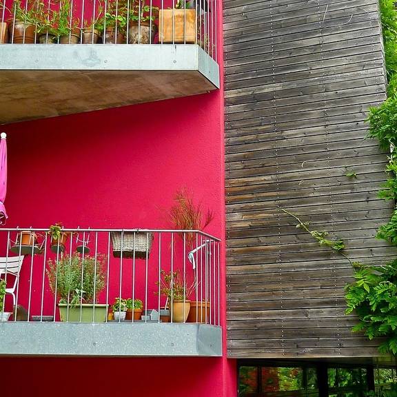 Ein Rotes Holz mit Holzfassade. Man sieht Balkone, auf denen grüne Pflanzen stehen.