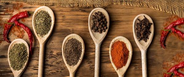 Holzkochlöffel, die mit unterschiedlichen Gewürzen gefüllt sind liegen geordnet auf einem Holztisch. Chilischoten liegen darum herum.