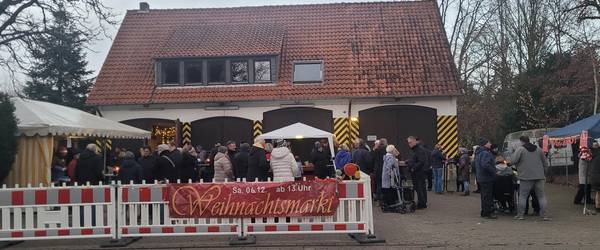 Blick auf das Gelände der alten Feuerwache Gleidingen. Im Vordergrund sind Menschen und Stände hinter einer Absperrung mit dem Banner "Weihnachtsmarkt" zu sehen. Im Hintergrund befindet sich das Gebäude der alten Feuerwache Gleidingen.