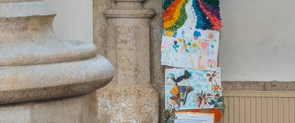 Sympolisches Kreuz aus bunt gemalten Kinderzeichnungen in einer Kirche. Eine Steinsäule auf der linken Seite, rechts eine Sitzbank mit Gesangsbuch.