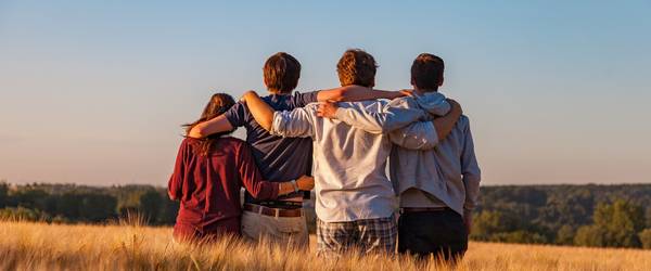 Vier Jugendlichen von hinten fotografiert, sitzen arm in arm auf einem Feld