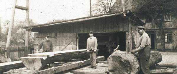 Drei Männer arbeiten mit Holz. Vor einem der Männer liegt ein großer Baumstamm, zwei anderer stehen neben einem gesägten Brett.